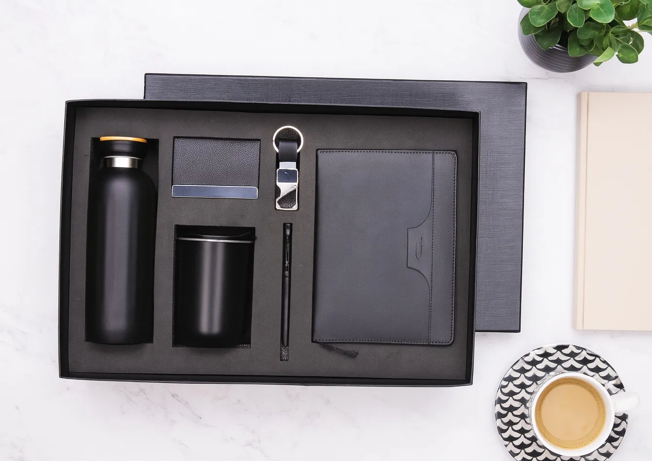 Open black gift box with insulated bottle, coffee tumbler, card holder, keychain, pen and leatherette diary on a marble surface.