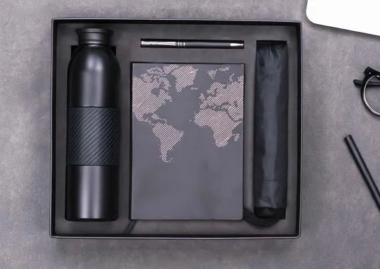 Black gift tray with a gripped water bottle, world-map diary, metal pen and folding umbrella on a grey surface