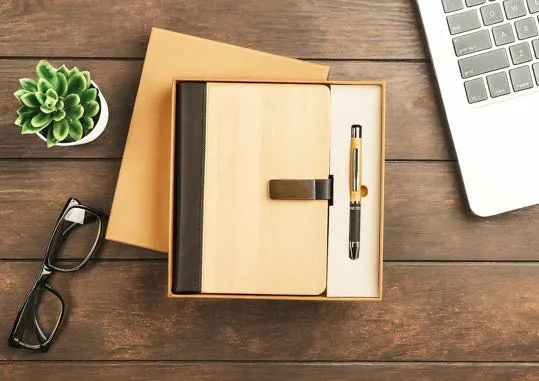 Bamboo-finish diary with metal clip and bamboo pen in a kraft box on a dark wood desk.