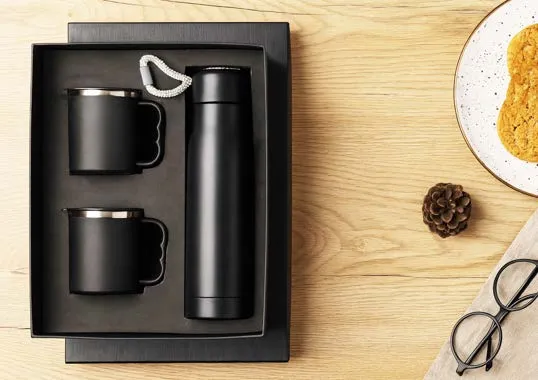 Gift box with two black steel mugs and a black flask with temperature-display cap on a wooden surface.