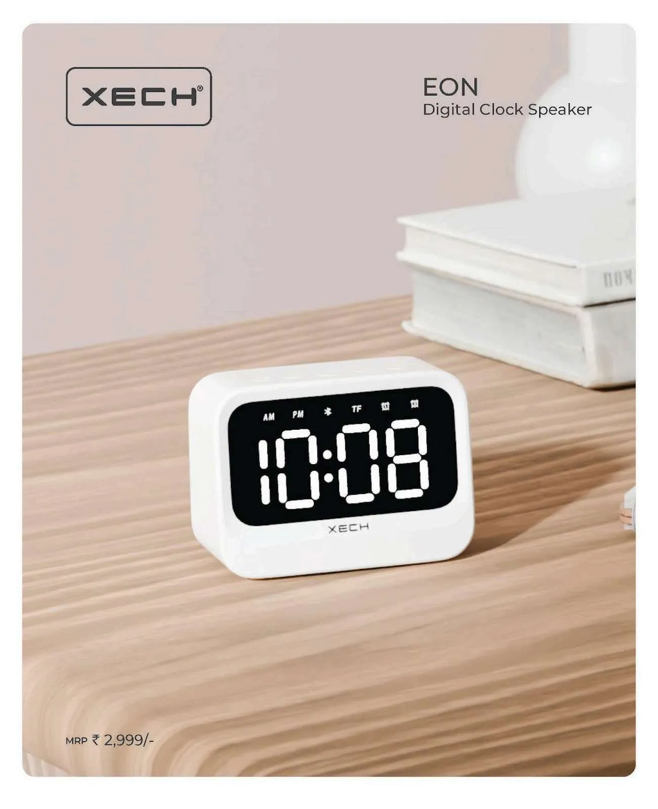 Compact white XECH Eon digital clock speaker showing 10:08 on a white LED display, on a wooden desk beside stacked books.