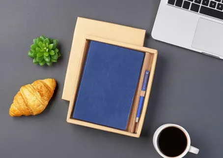 Blue leatherette diary with matching pen in a bamboo tray on a grey desk, beside coffee and a croissant.