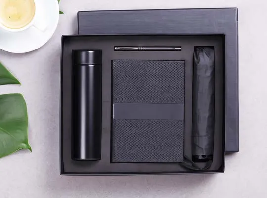 Black gift box holding insulated flask, textured diary, metal pen and folding umbrella beside a coffee cup