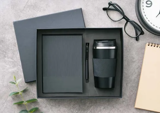 Black gift box with ribbed diary, slim pen and ribbed travel mug, beside glasses and a clock.