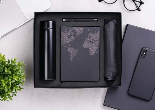 Black gift tray with insulated flask, world-map diary, metal pen and folding umbrella on a marble desk