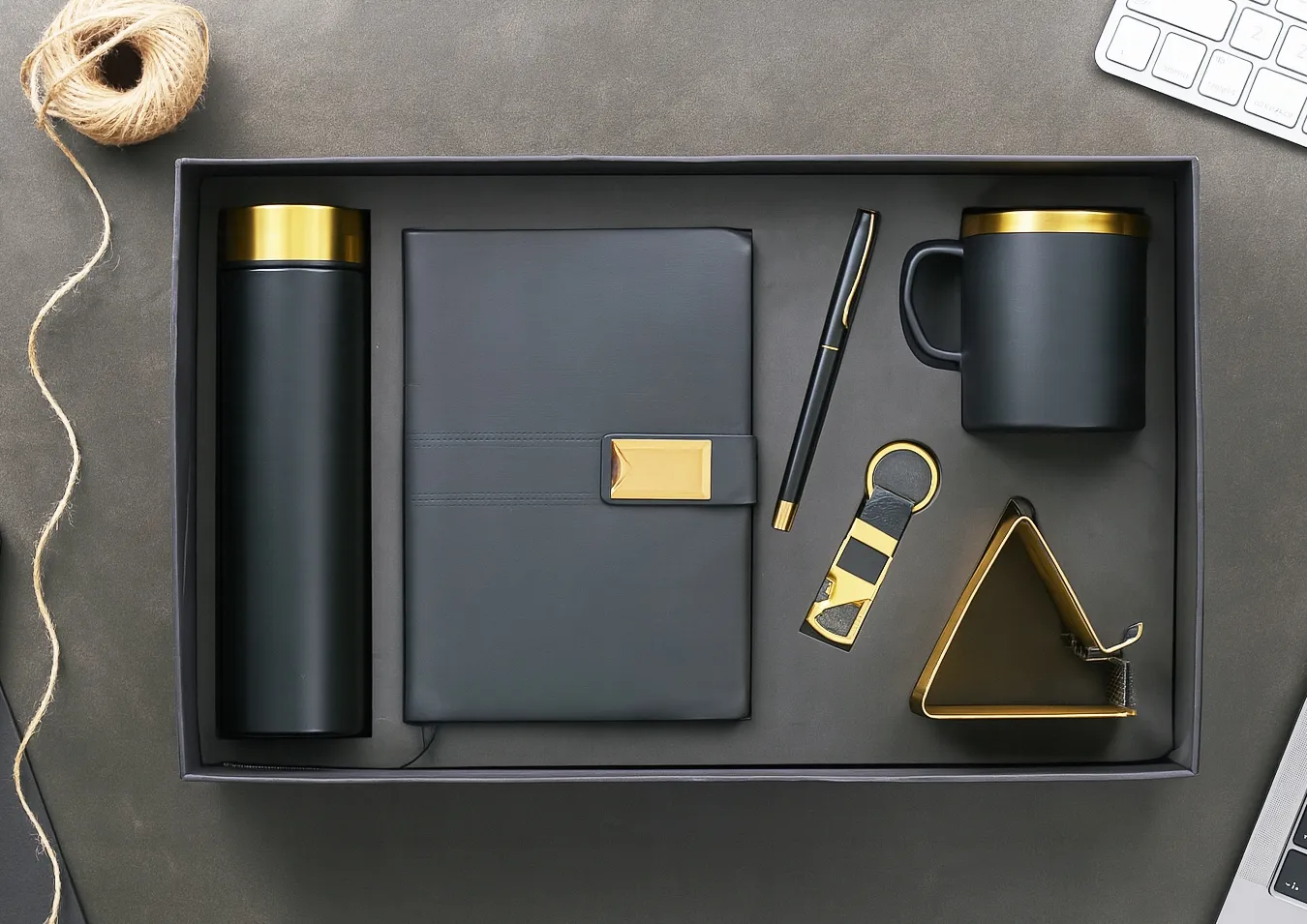 Open gift box with black-and-gold flask, grey diary, pen, keychain, triangular phone stand and mug on a dark surface