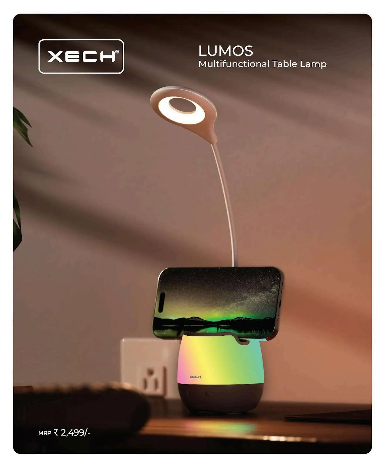 White XECH Lumos table lamp with glowing colour base holding a phone, on a desk near a wall socket.