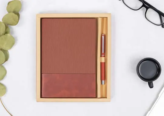 Tan ribbed diary with a wooden pen in a bamboo tray on a white desk, beside eucalyptus and a black mug.