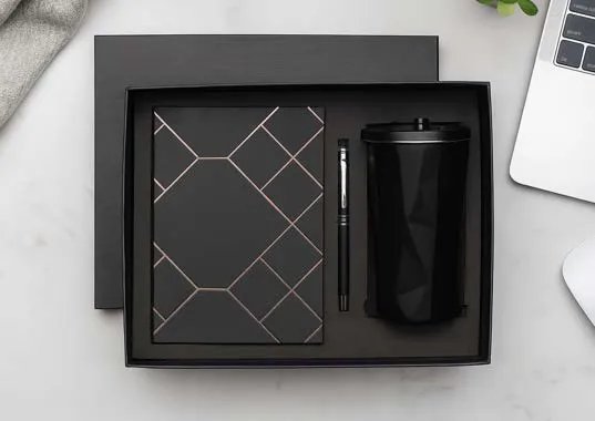 Black gift box holding a diamond-pattern diary, a pen and a black lidded tumbler beside a laptop.