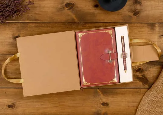 Open kraft box with a cognac leatherette diary featuring a gold border and buckle, beside a metal pen on wood