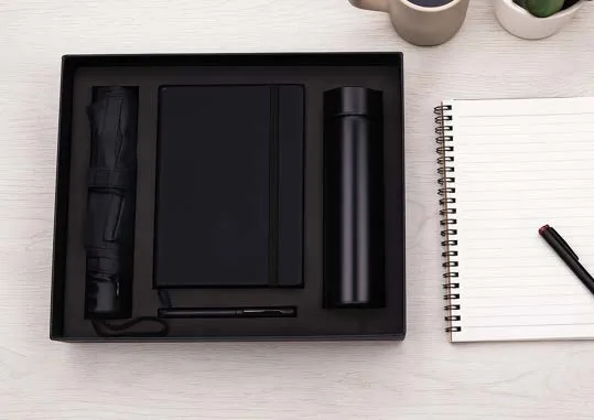 Black gift box with folding umbrella, A5 diary, vacuum flask and pen on a pale wood desk.