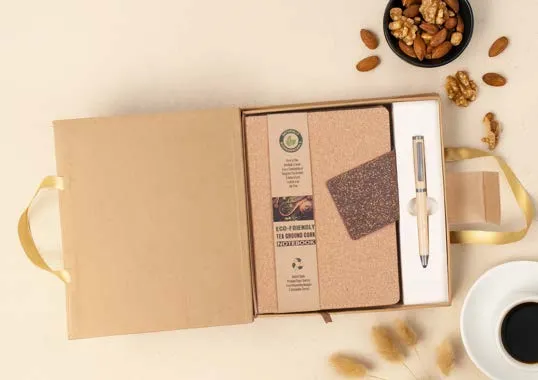 Open kraft gift box holding a cork notebook and a slim metal pen on a cream surface