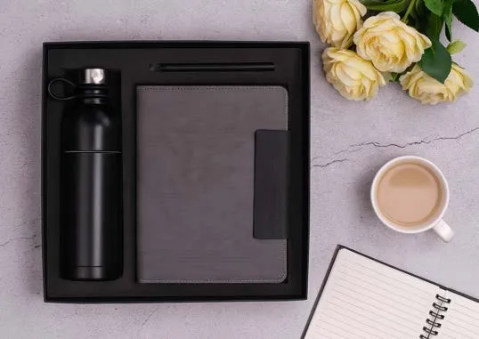 Gift box with black bottle, grey flap diary and pen, styled with yellow roses and a cup of tea.
