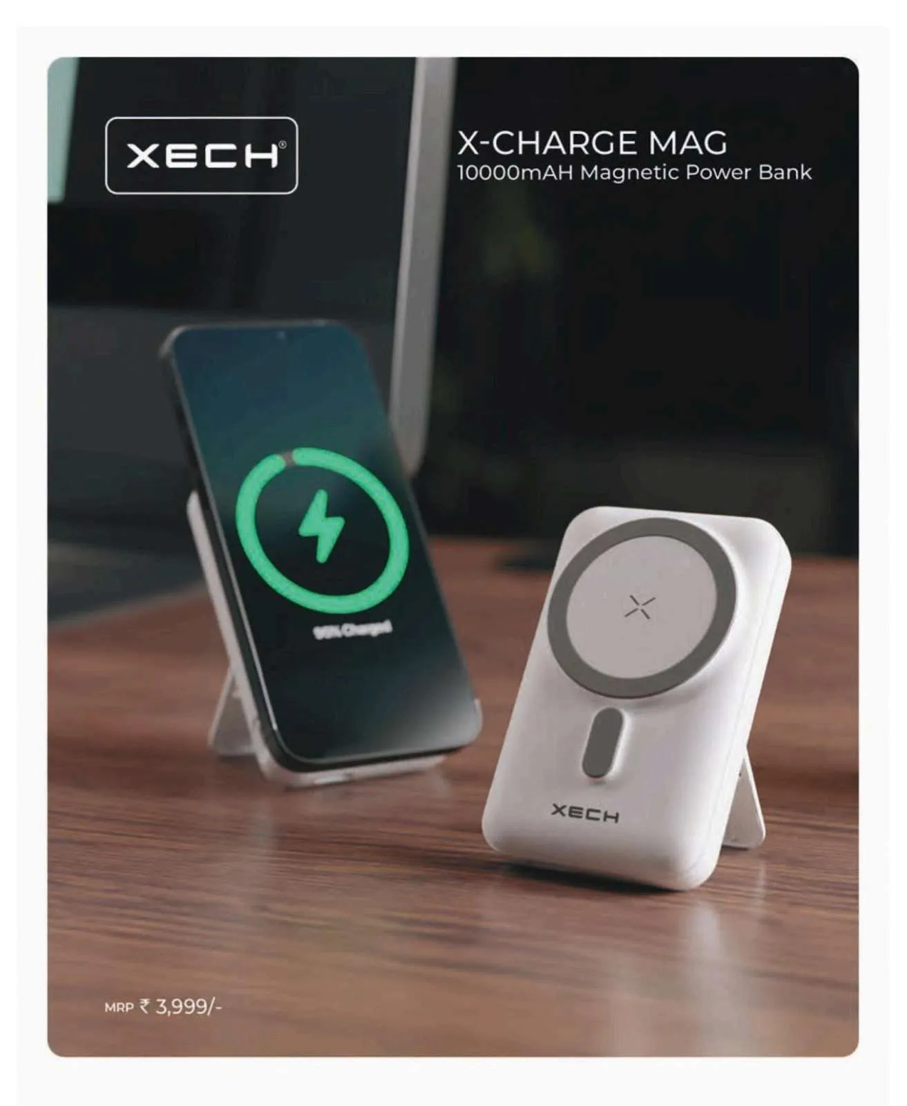 White XECH X-Charge Mag magnetic power bank with kickstand beside a phone wirelessly charging on a wooden desk.